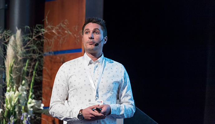 En man i vit skjorta med mönster står på en scen, med en blombukett och en träpanel i bakgrunden.