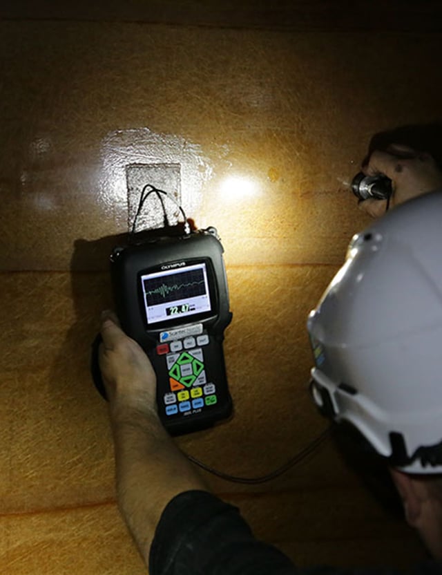 En person i hjälm använder en ultraljudsdetektor för att undersöka en yta, med en ficklampa som lyser upp området.