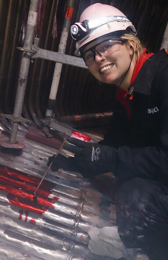 En person arbetar med att applicera röd färg eller markering på metallrör i en industriell miljö, med skyddsutrustning inklusive hjälm och handskar.