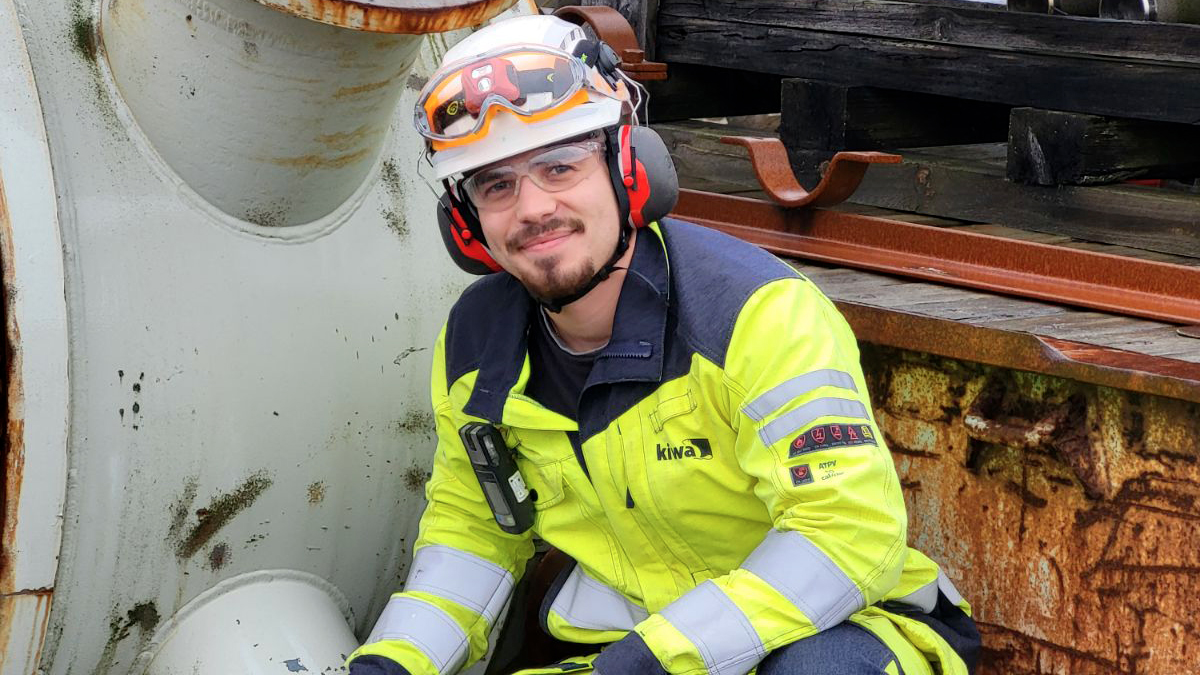 Person som bär gul skyddsklädsel, reflexband, hjälm med hörselskydd och skyddsglasögon, arbetar vid industriell utrustning med rostiga metalldelar i bakgrunden.