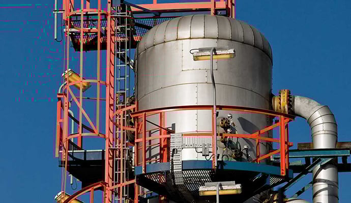 Industriell anläggning med en stålcistern omgiven av röda och blå metallramar och stegar under klarblå himmel.
