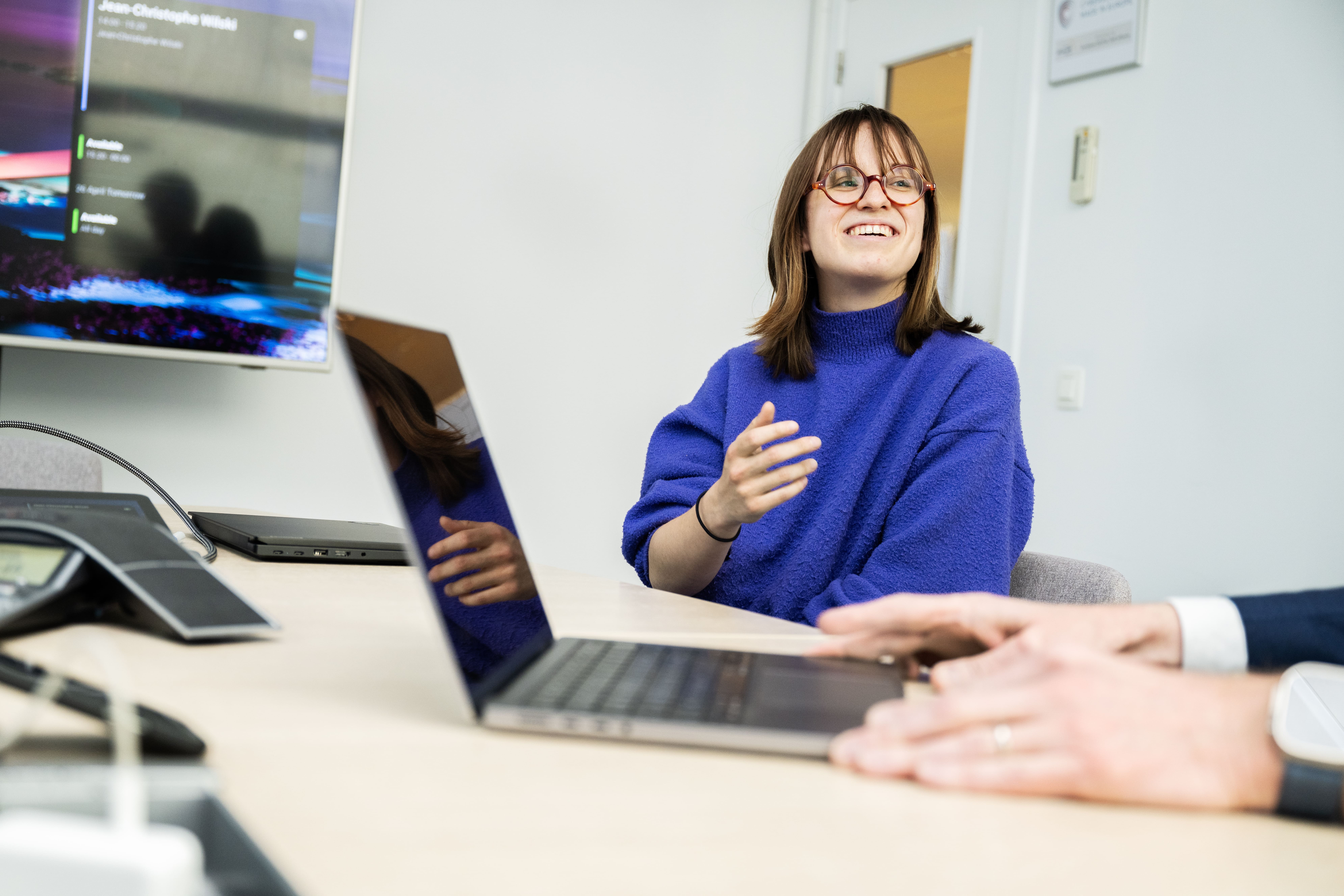 A woman in a blue sweater with glasses is smiling and engaged in conversation during a business meeting. She is sitting at a conference table with laptops and a large screen displaying content in the background