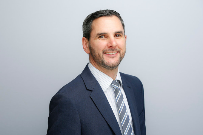 A man in a navy blue suit, striped tie, and collared shirt, standing against a light gray background.