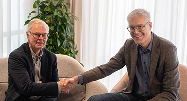 Två personer sitter på soffor och skakar hand, med en grön växt och ljusa gardiner i bakgrunden.