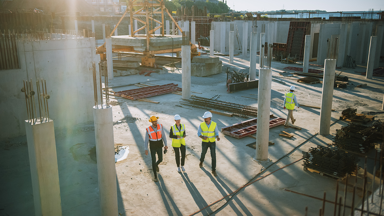 Construction site with four Kiwa auditors in safety gear examining building progress under sunlight, surrounded by concrete columns and construction materials