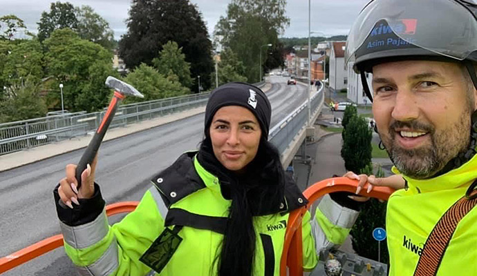 Personer i arbetskläder och skyddsutrustning står i en skylift vid en väg, omgivna av grönska och byggnader.
