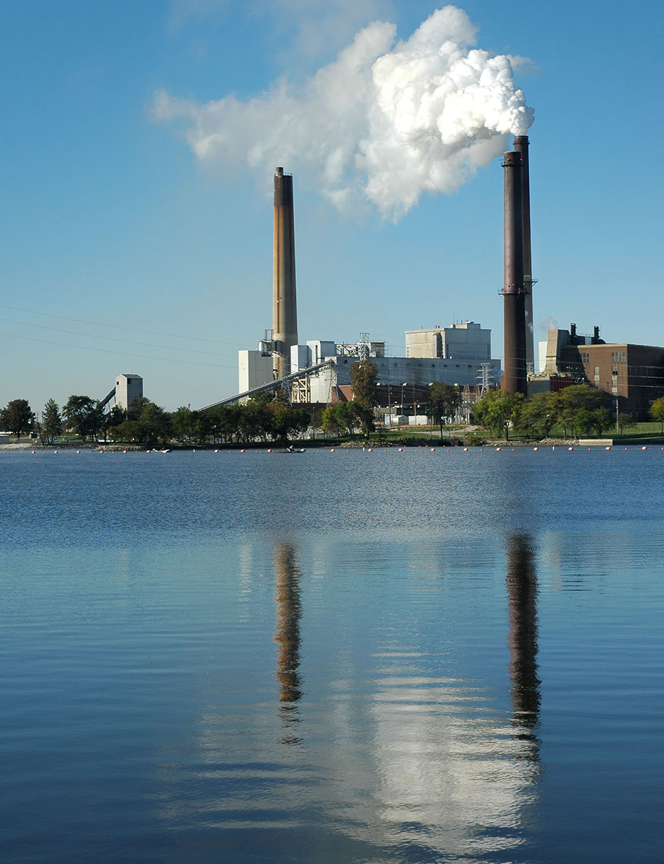 Kraftverk med två skorstenar som släpper ut vit rök, beläget vid vattnet med spegelbild i det blå vattnet.