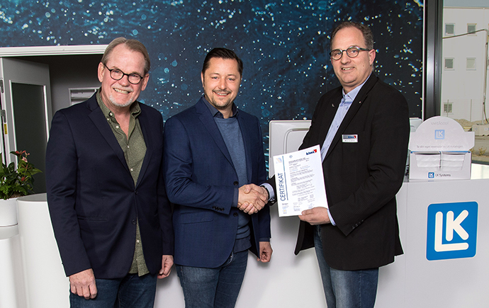 Tre personer står vid en receptionsdisk; två håller ett handslag medan en annan håller ett papper. Bakgrunden har en modern design med en företagslogotyp och byggnadsstruktur.