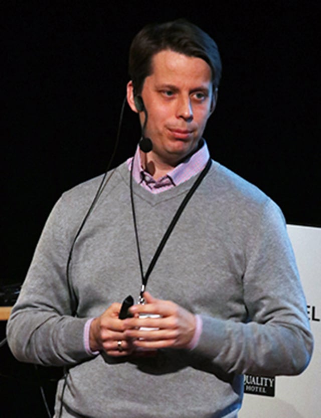 Bj&ouml;rn Lindberg fr&aring;n Arbetsmilj&ouml;verket, i gr&aring; tr&ouml;ja och rosa skjorta, h&aring;ller en presentation med mikrofon och fj&auml;rrkontroll. Bakgrunden visar en svart scen och en vit skylt med text.