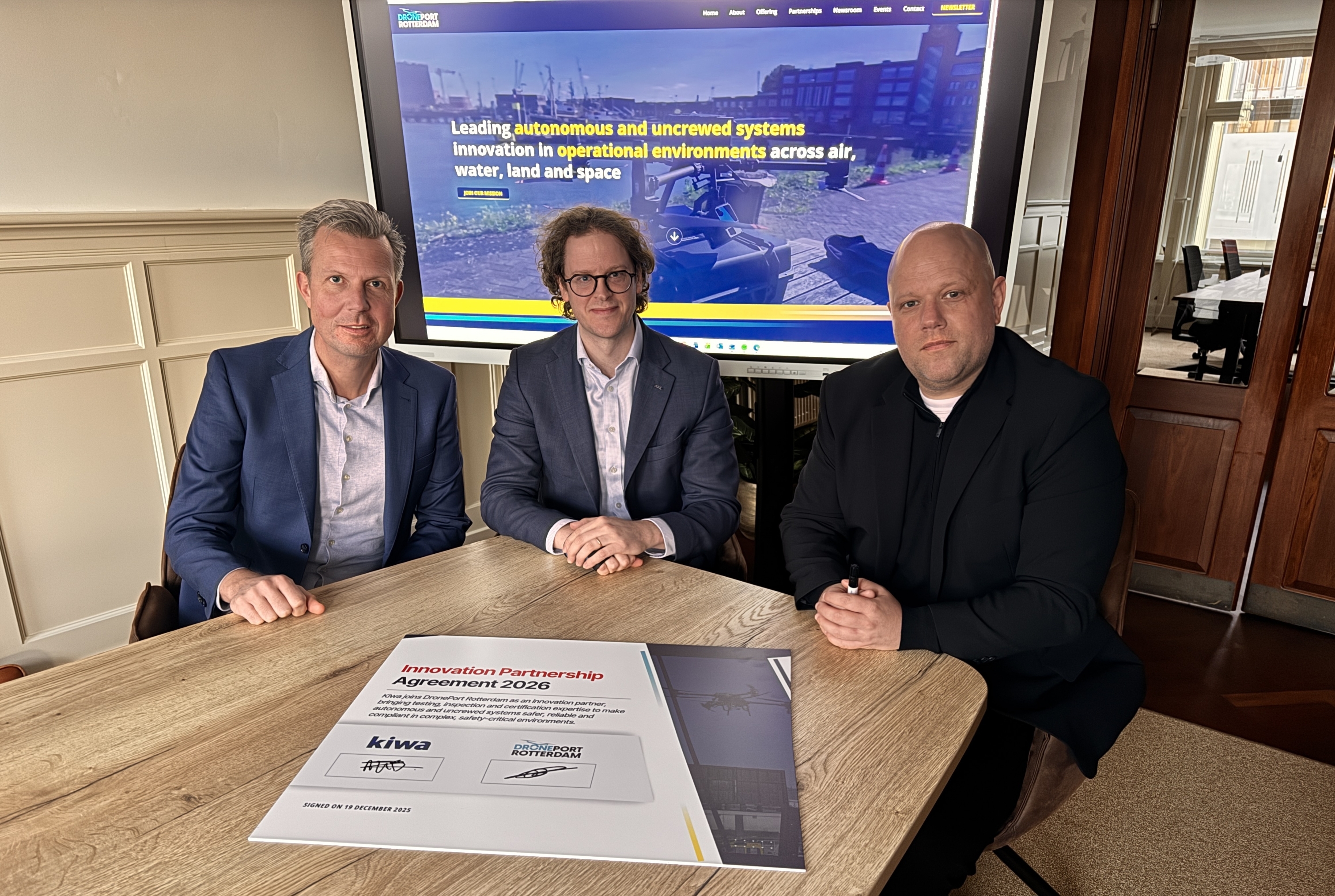 Three individuals seated at a table with a document featuring logos, in front of a screen displaying a graphic and text about autonomous and unmanned systems innovation in operational environments.
