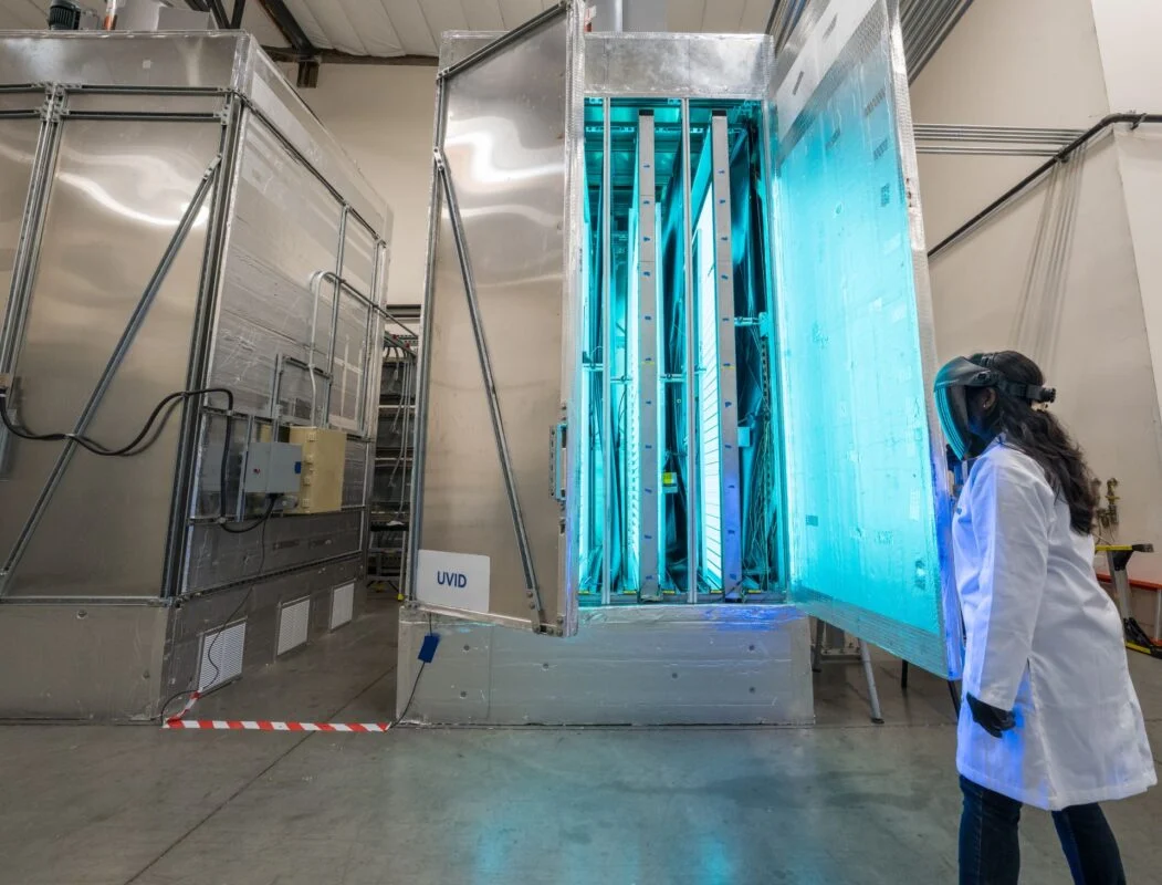 A Kiwa lab employee in protective gear examines a large, illuminated UV sanitation chamber in a laboratory setting