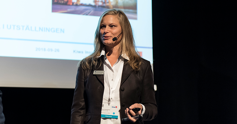 En person i mörk kavaj och vit skjorta står framför en skärmpresentation, med en mikrofon eller fjärrkontroll i handen.