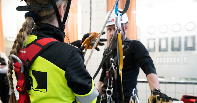 Personer som utför säkerhetsarbete på höjd med sele, rep och skyddsutrustning i en industriell miljö.