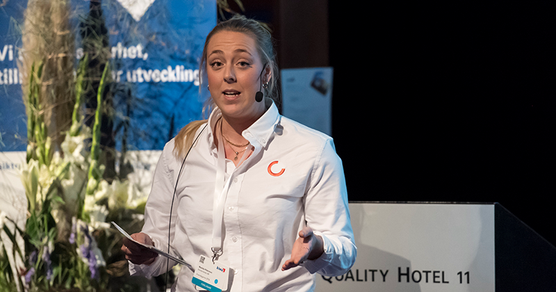 Kvinna i vit skjorta står och presenterar med en namnbricka runt halsen, blommor och en blå bakgrund synliga i miljön.