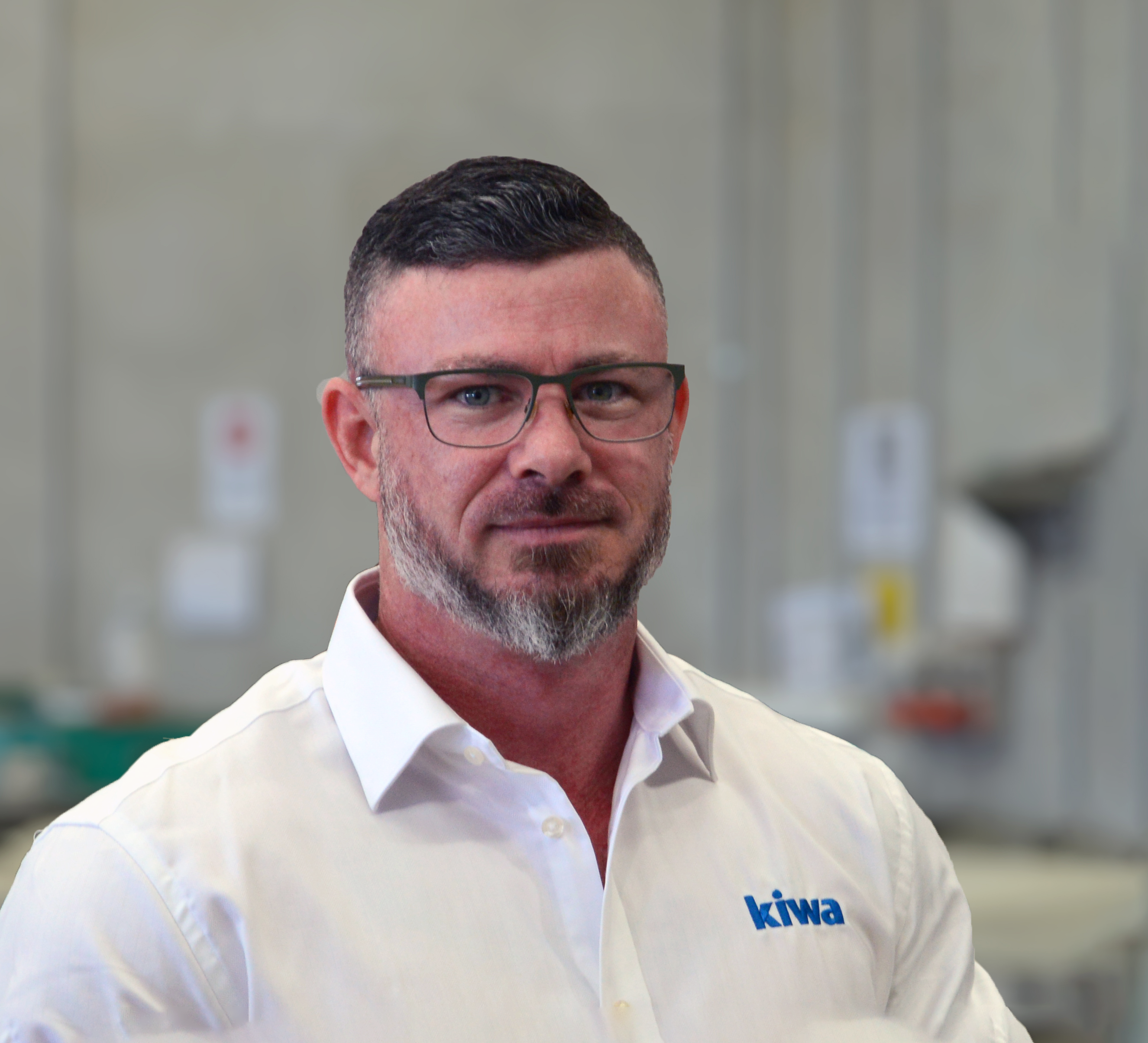 Kiwa employee wearing glasses and a white shirt with the "kiwa" logo, standing in an industrial setting