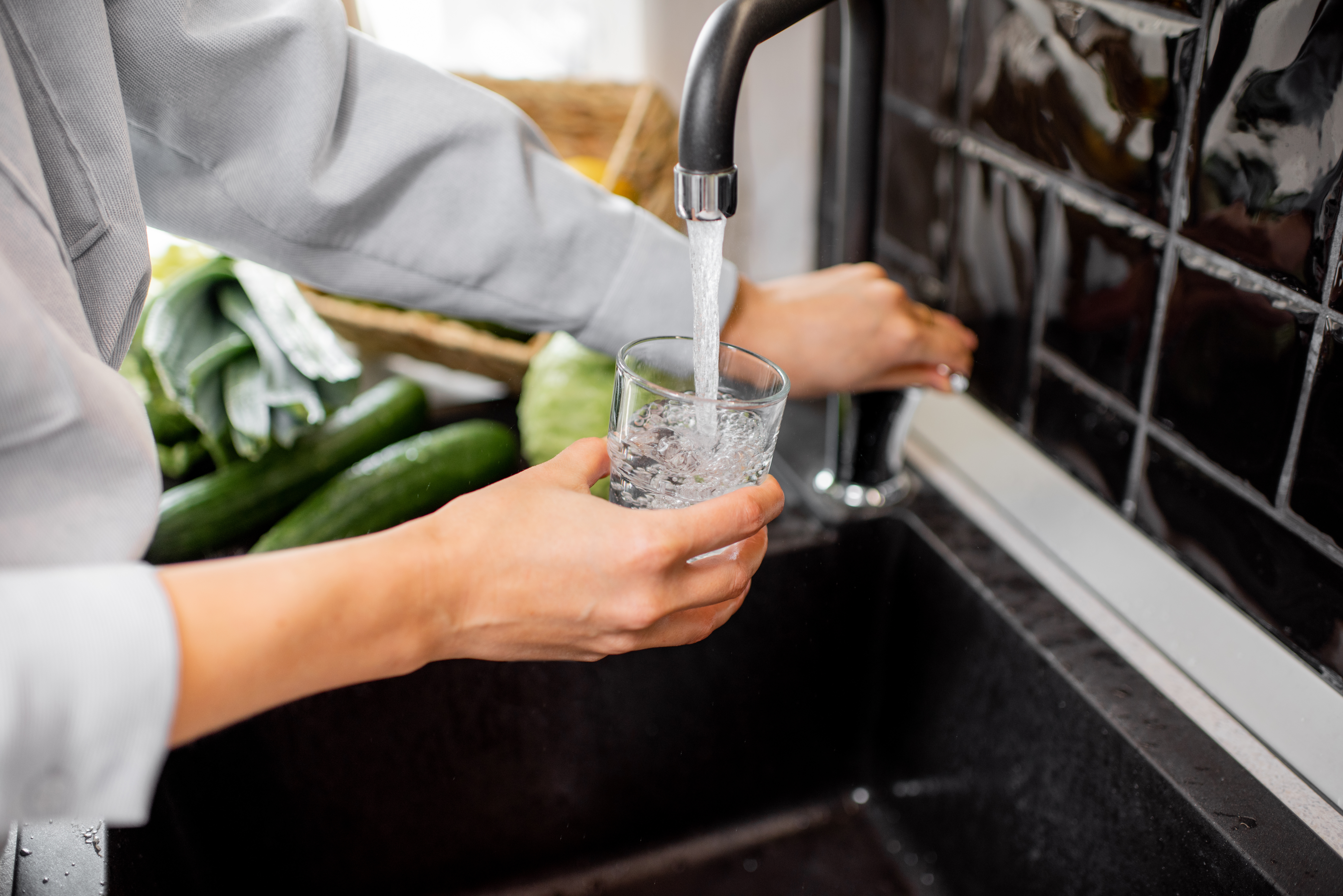 Person filling a glass with water from a kitchen faucet, surrounded by fresh green vegetables
