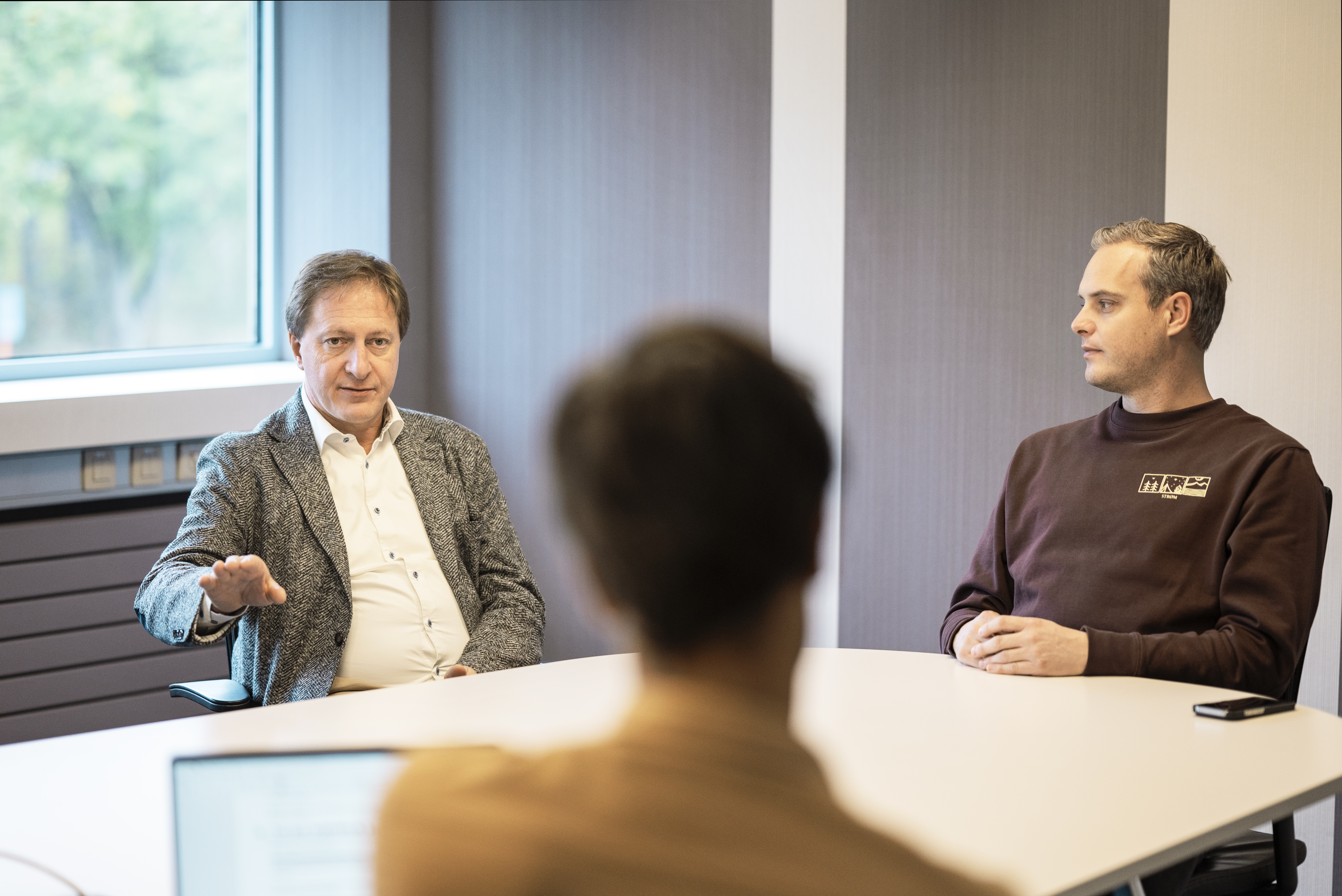 Three people in a modern office setting having a discussion around a conference table, with a window in the background