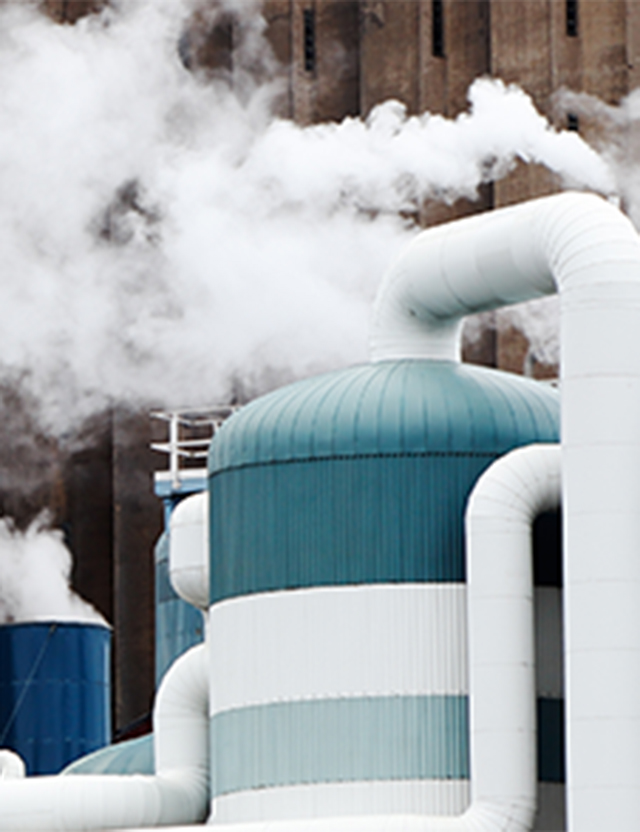 Industribyggnad med stora rör och tankar som släpper ut vita ångor mot en bakgrund av tegelväggar.