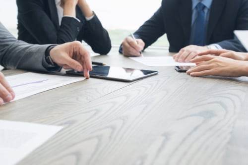 Personas reunidas en una mesa de madera, revisando documentos y usando dispositivos electrónicos mientras llevan ropa formal.