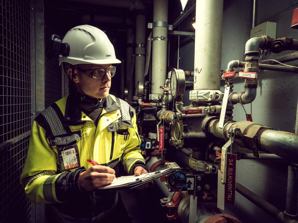 Kiwa engineer in safety gear inspecting industrial pipes and gauges, taking notes on a clipboard in a mechanical room