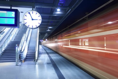 Tågplattform med en röd tunnelbana som rör sig snabbt, en klocka och en trappa till vänster.