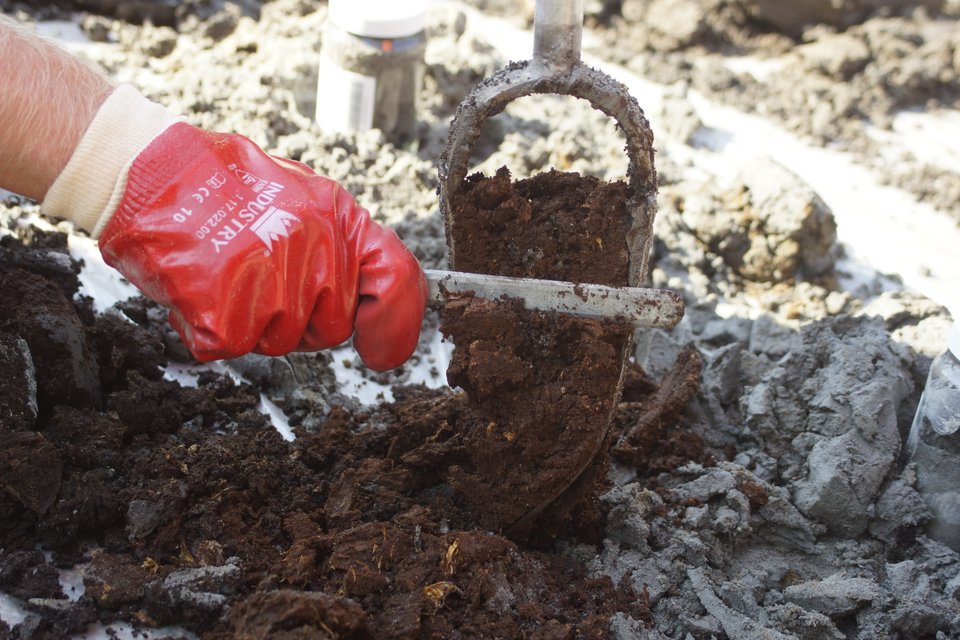 Hand met rode handschoen gebruikt een grondboor om aarde te verzamelen, omringd door grond en steenachtige materialen.
