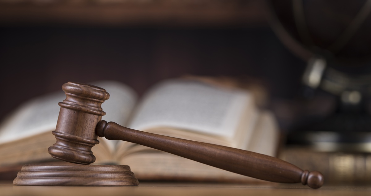 Houten hamer van een rechter op een houten onderstel, met een geopende wetboek en globes vaag op de achtergrond.