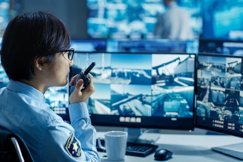 Beveiligingsmedewerker in uniform met een portofoon, werkend aan een bureau met meerdere monitorschermen waarop beveiligingsbeelden te zien zijn.