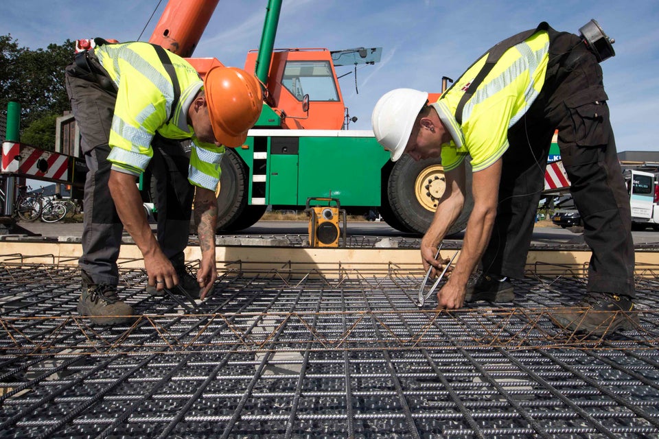 Twee bouwvakkers werken aan een fundering met stalen vlechtwerk en betonijzer, met een kraanwagen op de achtergrond.