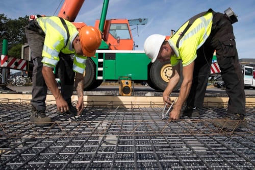 Twee bouwvakkers werken aan een fundering met stalen vlechtwerk en betonijzer, met een kraanwagen op de achtergrond.