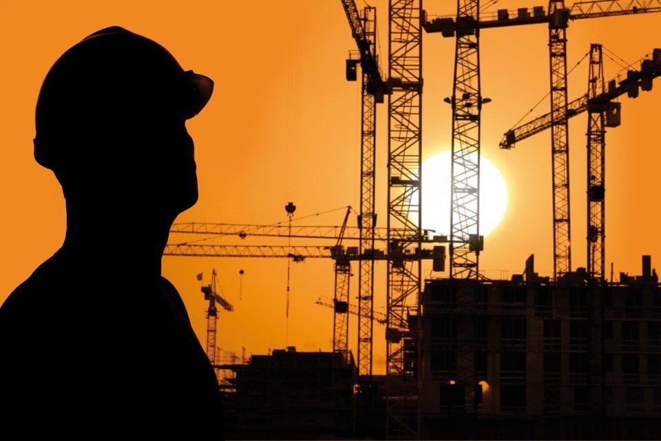 Silhouet van een bouwvakker met helm, tegen een achtergrond van hijskranen en een oranje zonsondergang.