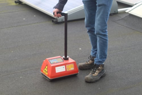 Persoon gebruikt een draagbaar apparaat met een rode behuizing en waarschuwingssymbolen op een donkere vloer, mogelijk op een plaats met zonnepanelen.