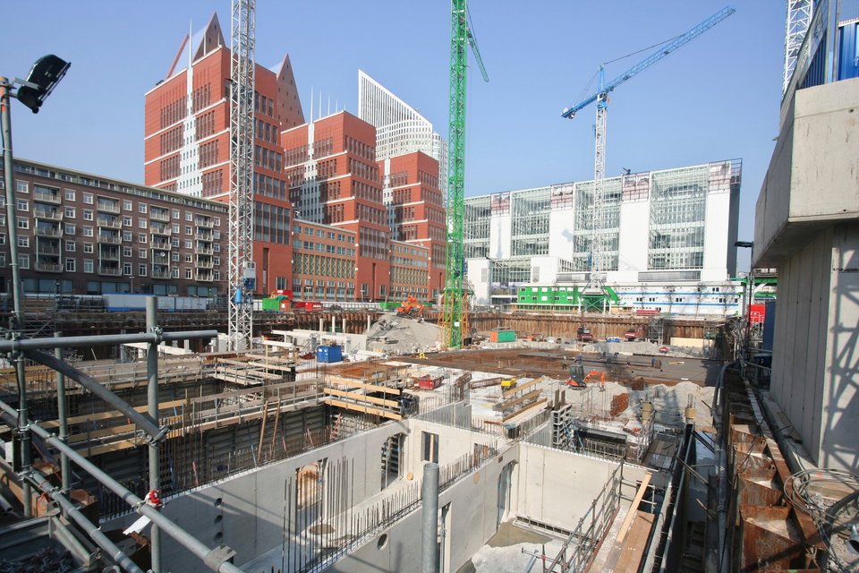 Bouwplaats met graafwerkzaamheden, omringd door moderne gebouwen en hijskranen in een stedelijke omgeving.
