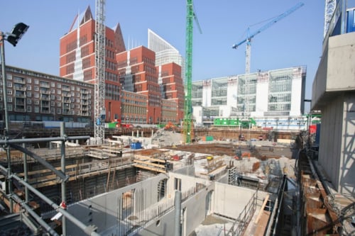 Bouwplaats met graafwerkzaamheden, omringd door moderne gebouwen en hijskranen in een stedelijke omgeving.