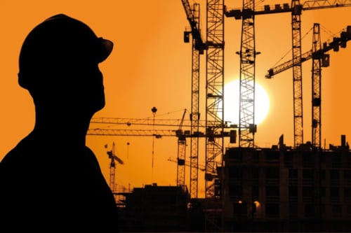 Silhouet van een persoon met een bouwhelm voor een achtergrond van hijskranen en gebouwen bij zonsondergang.