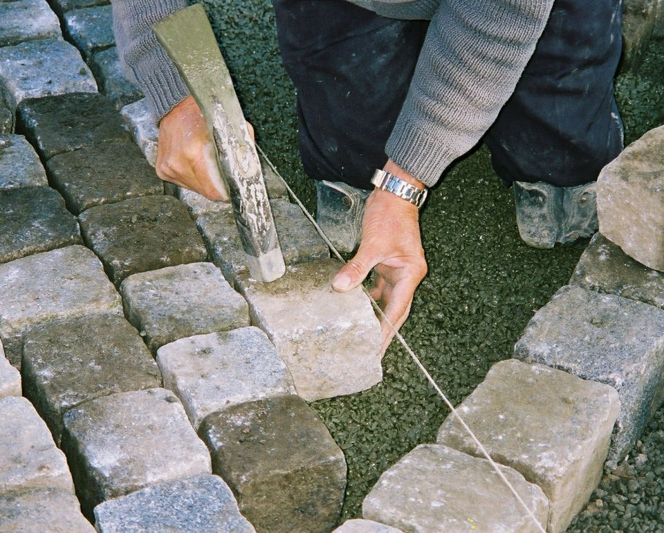 Close-up van handen die straatstenen bewerken met een hamer, op een ondergrond van grind.