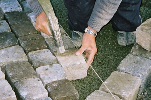 Close-up van handen die straatstenen bewerken met een hamer, op een ondergrond van grind.