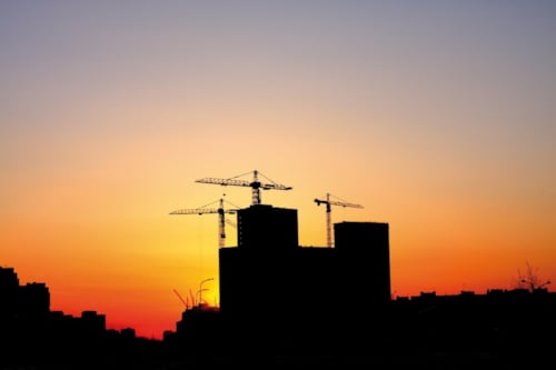 Silhouet van bouwkranen en gebouwen tegen een kleurrijke zonsondergang.