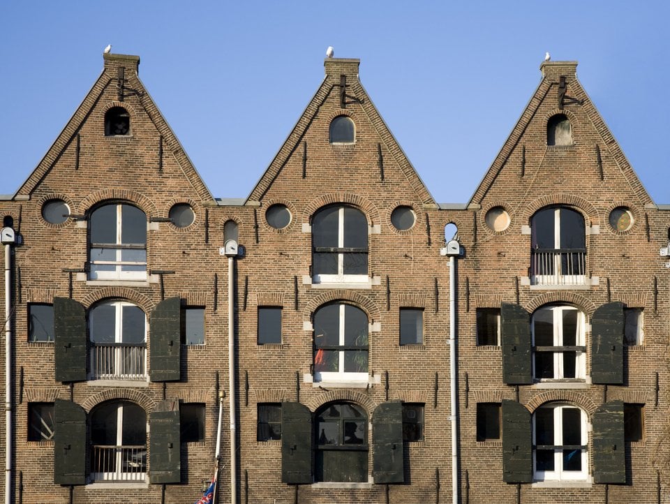 Bakstenen gebouw met drie puntgevels, groene luiken en ronde ramen, gefotografeerd tegen een blauwe lucht.