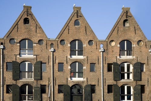 Een rij driehoekige gevels van een historische bakstenen gebouw met donkere houten luiken en ramen, tegen een blauwe lucht.