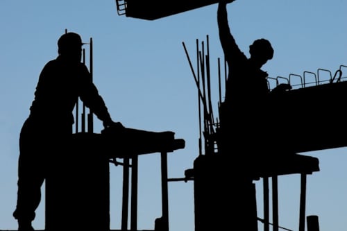 Silhouetten van bouwvakkers die werken op een bouwplaats tegen een blauwe lucht.