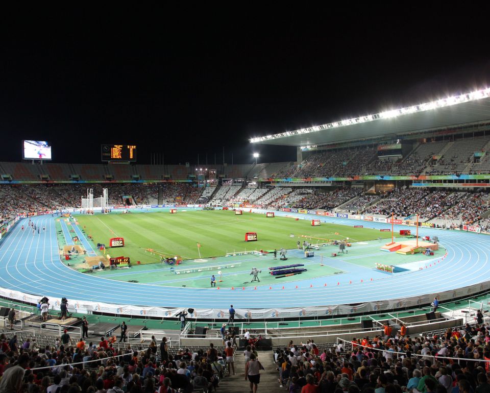 Atletiekstadion bij nacht, gevuld met toeschouwers, een blauwe atletiekbaan en verschillende sportarena's op het veld.