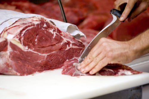 Een persoon snijdt rauw rood vlees op een witte snijplank met een mes.