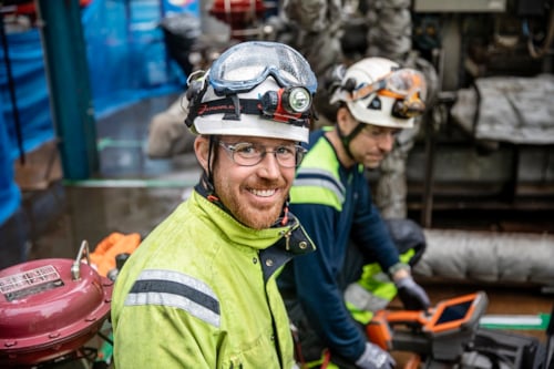 Two workers wearing high-visibility jackets and protective helmets with lights, operating equipment in an industrial setting.