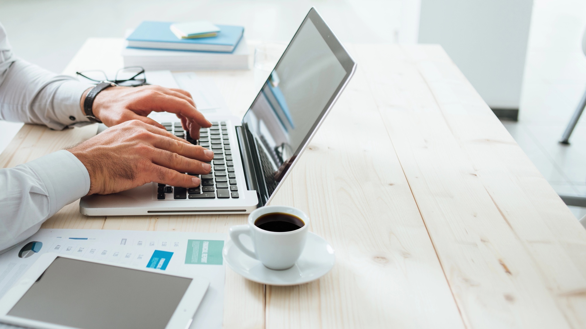 Man typing on a laptop at a wooden desk with coffee cup, tablet, and documents nearby.