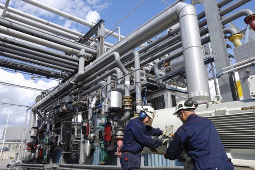 Twee technici in werkkleding inspecteren industriële leidingen en installaties op een fabrieksterrein.