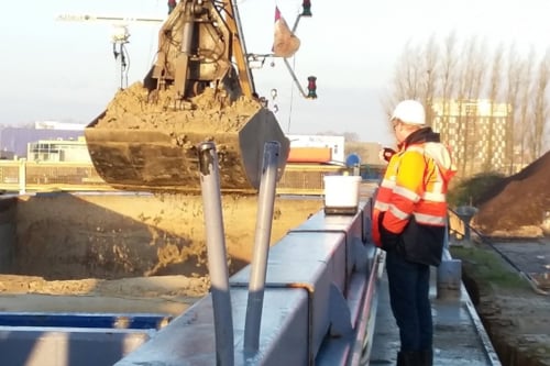 Man in een oranje veiligheidsjas en witte helm observeert een graafmachine die zand verplaatst op een bouwplaats.