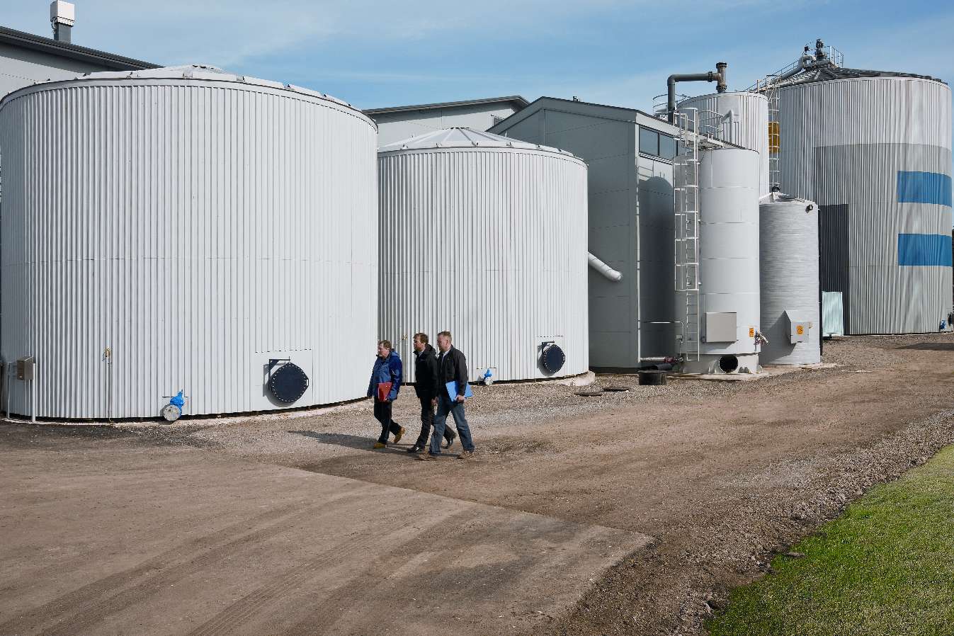 Flera stora, cylindriska tankar av metall utomhus på en industriell anläggning, med tre personer som går längs en grusväg i förgrunden.