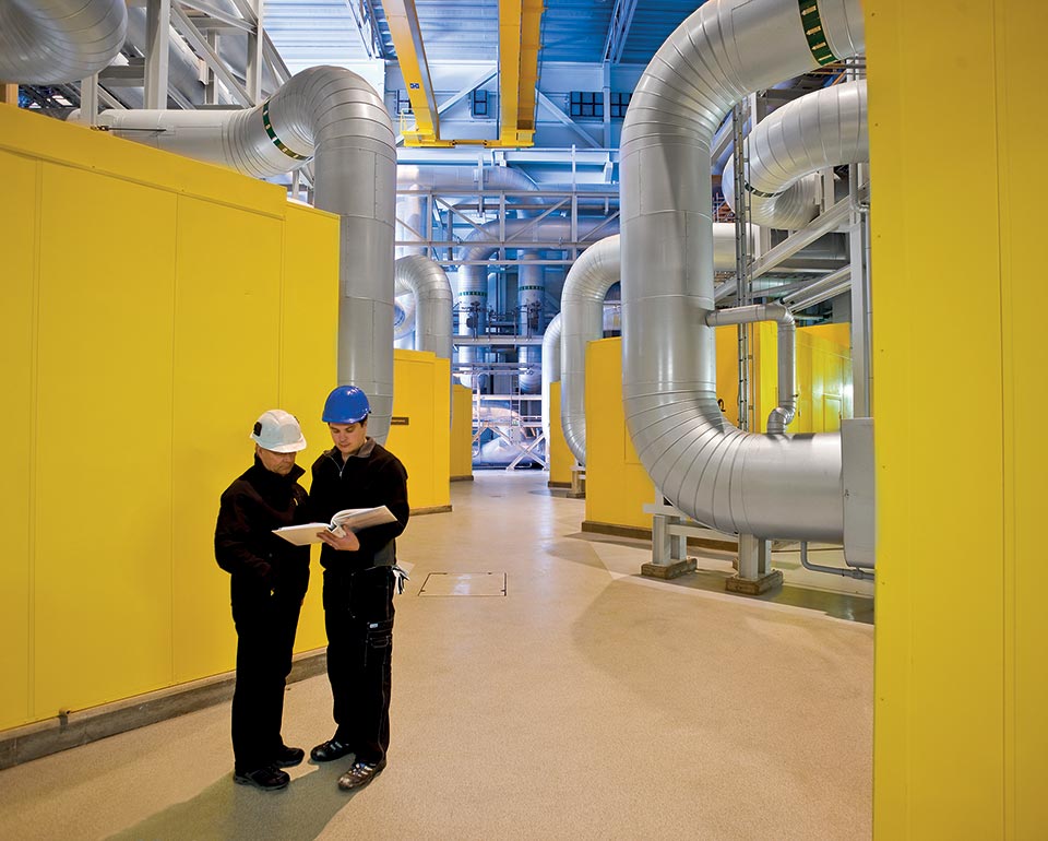 Två personer i arbetskläder och hjälmar står och håller dokument i en industriell anläggning med stora rör och trycksatta anordningar med gula väggar. 