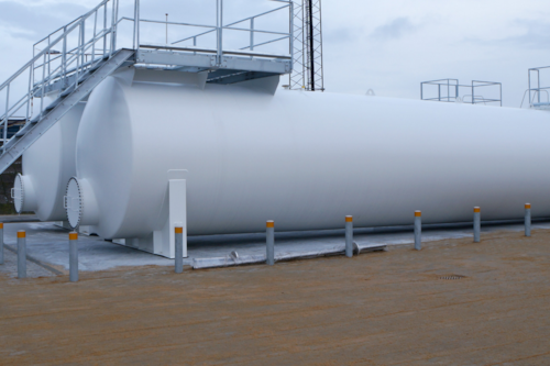 Vit cylindrisk tank utomhus på en industriell plats, omgiven av stolpar och med en trappa som leder upp till toppen.